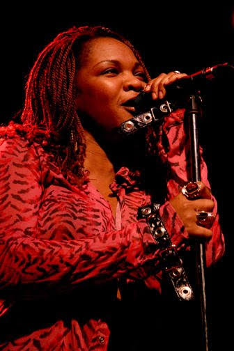 Trina Meade singing close to the microphone in a red patterned shirt under deep red stage light