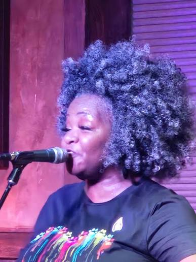 Trina Meade singing into a handheld microphone in a small club, lit magenta