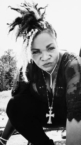 Black-and-white full-length portrait of Trina Meade crouched outdoors in a pine clearing